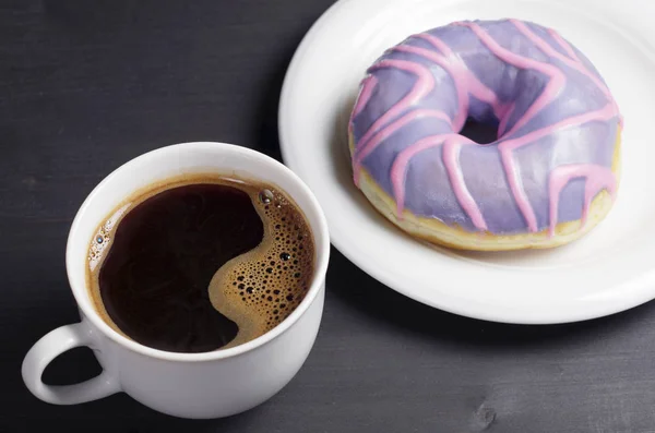 Cup of coffee and a donut with a violet glaze on blact wooden table close up