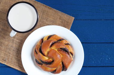 Emaye bir fincan süt ve mavi ahşap masa, üstten görünüm üzerinde beyaz tabak içinde çikolata kremalı tatlı çörek