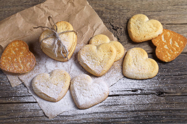 Shortbread cookies in shape of heart tied with a rope and several separate ones sprinkled with powdered sugar on paper located on a dark wooden surface