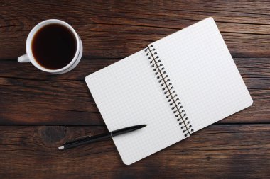Bir fincan kahve ve ahşap arka planda kalemle açılmış bir defter. Üst görünüm