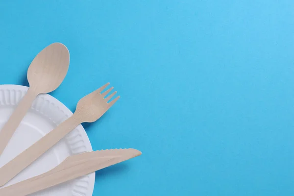 Wooden cutlery on a disposable paper plate on blue background, top view with copy space