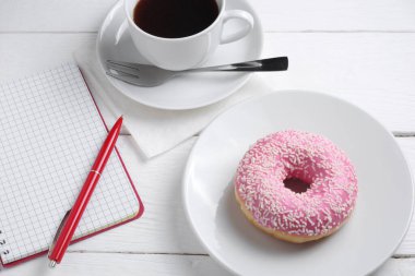 Not defteri ve beyaz ahşap masada kahve fincanı olan pembe çörek.