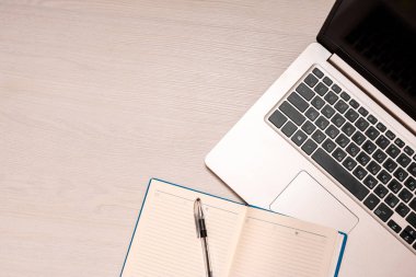 Beyaz ahşap masa üzerinde siyah kalem ve gümüş dizüstü bilgisayar ile açılan defter, üst görünüm, spazm kopya