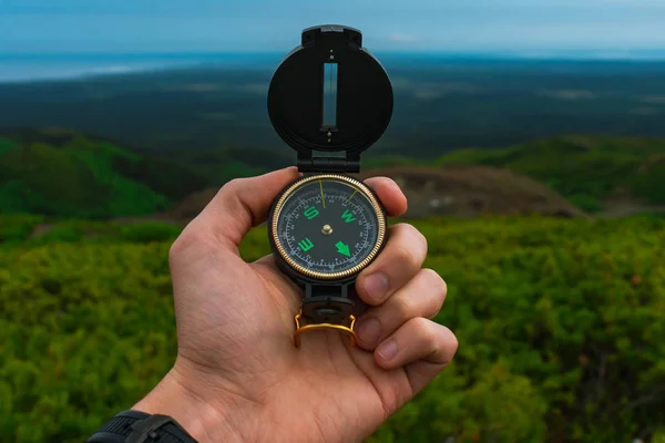 Travel, camping, orienteering and navigation concept - black magnetic compass close-up in a man s hand, blurred landscape background