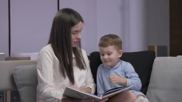 Mutter und Sohn lesen ein Buch sitzend auf einem Sofa — Stockvideo