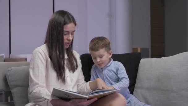 Mutter und Sohn lesen ein Buch sitzend auf einem Sofa — Stockvideo