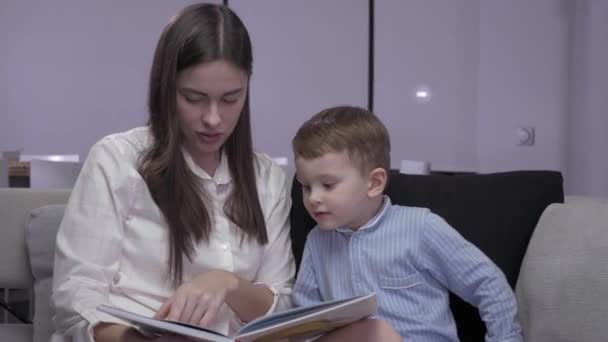 Mom and son are reading a book sitting on a sofa — Stock Video