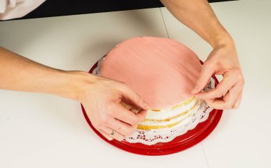 Kırmızı plastik tepside katmanlı kekin üzerine pembe badem ezmesi ekleyen kişi.