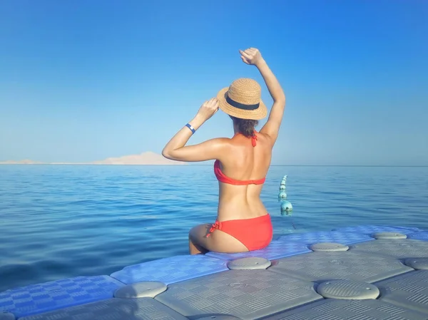 Bikini ve şapka mavi deniz ve parlak gökyüzü önünde kayan plastik duba üzerinde oturan genç kadın