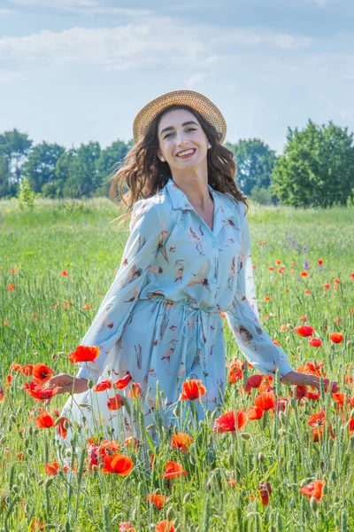 Kırmızı haşhaş tarlasında gülümseyen açık mavi elbiseli ve hasır şapkalı genç kadın.
