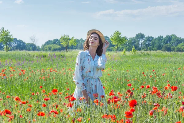 Açık mavi elbiseli ve hasır şapkalı genç bir kadın kırmızı haşhaş tarlasında gözleri kapalı duruyor.