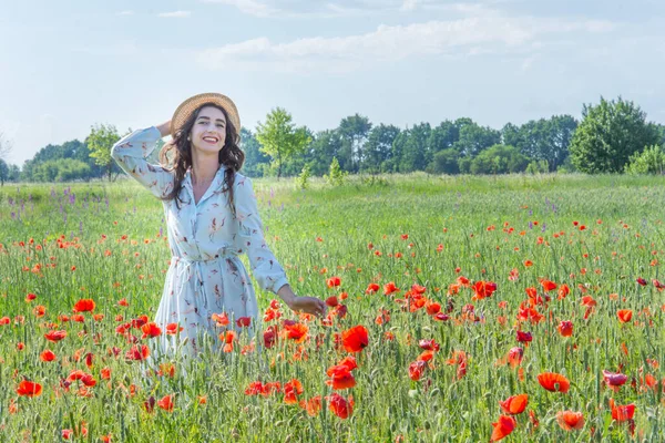 Kırmızı haşhaş tarlasında gülümseyen açık mavi elbiseli ve hasır şapkalı genç kadın. 