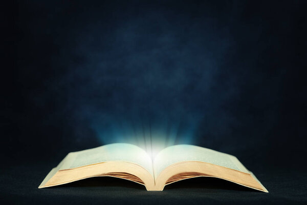 Open magic book emitting light on dark textured background and copy space