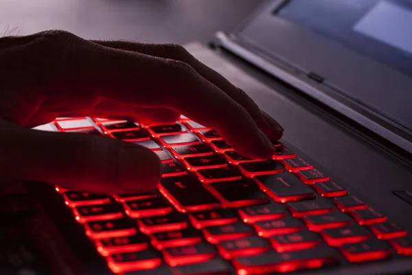 Close up of fingers typing on glowing red backlight keys on a computer keyboard. in the dark. Computer hacker, internet fraud or spying concept.