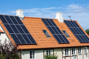 Mavi arka planı olan eski bir evin kırmızı kiremit çatısındaki modern siyah güneş panelleri.