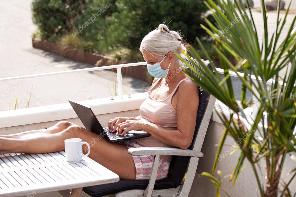 Sun tanned senior woman in shorts sitting on a balcony wearing a medical face mask to protect against Coronavirus or Covid-19 pandemic while working on a laptop computer. Working from home concept in modern white building environment.