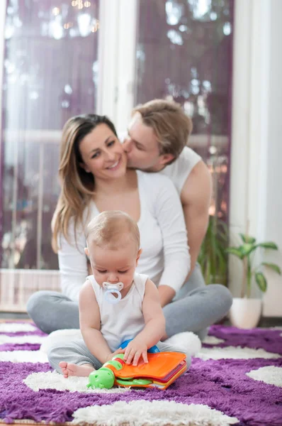 美しい小さな赤ちゃん床に座って おもちゃで遊んでいます バック グラウンドでの幸せな両親 母のキス — ストック写真