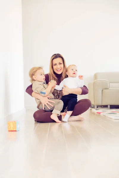 Feliz madre sentada en el suelo con sus dos bebés y jugando . —  Fotos de Stock