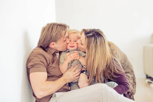幸せな家族が彼らの小さな赤ちゃんを床に座って。家族は彼らの息子と自宅で時間を過ごします。親は彼らの子供のキス. — ストック写真