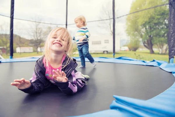 Schönes kleines Mädchen, das auf dem Trampolin liegt und posiert. Kleiner Junge springt in den Hintergrund. — Stockfoto