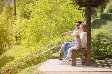 Baba, anne ve onların küçük kız bir güneşli bahar gününde tahta bankta oturan.