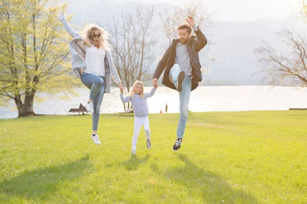 Buon padre, madre e bambina che saltano vicino al lago in una bella giornata di sole . — Foto Stock