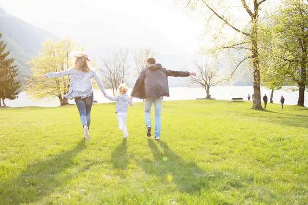 Buon padre, madre e bambina che corrono lungo il lago e godono in una bella giornata di sole . — Foto Stock