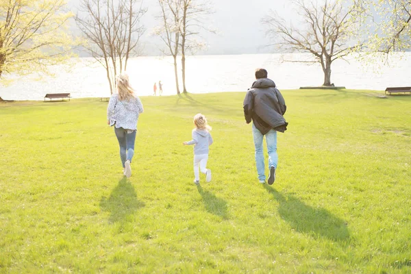 Buon padre, madre e bambina che corrono lungo il lago e godono in una bella giornata di sole . — Foto Stock