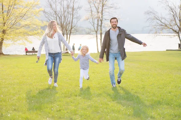 Buon padre, madre e bambina che corrono lungo il lago e godono in una bella giornata di sole . — Foto Stock