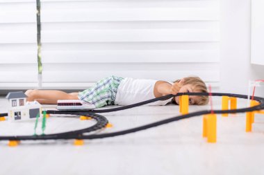 Sevimli çocuk yere uzanıp oyuncak demiryolu ile oynuyor.