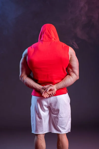 フード付きの強力な筋肉質の男が赤いスウェットシャツを着てポーズをとっている バックビュー スタジオ撮影で赤と青の煙 — ストック写真