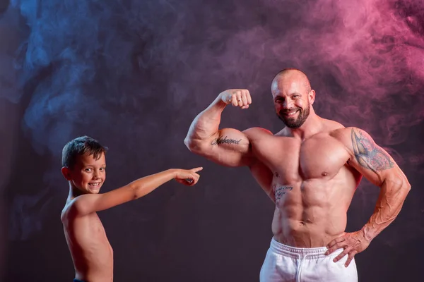 赤と青の煙とスタジオで彼の息子と一緒にポーズを取る強力な筋肉の男 両足の収縮を示す小さな男の子 — ストック写真