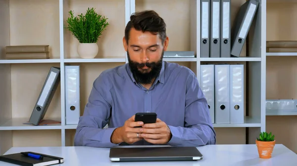 worker in start up company sitting in modern office using smart phone. young man with beard and mustache wearing blue shirt texting message or chatting online with friend. happy employer use mobile in workspace. hipster sitting at the desk in room wi
