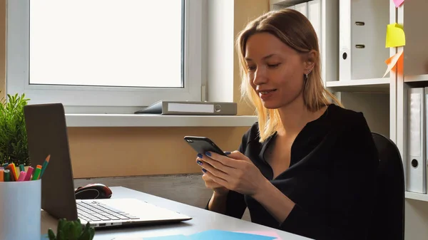 スマート フォン携帯メール メッセージを使用して大人の白人女性 モバイル幸せ笑顔のアプリを使用して黒のドレスを着ている金髪の女性 従業員ウィンドウで小さい事務所で机に座って — ストック写真