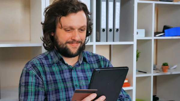 hipster man with beard pay bills online using app on digital tablet. handsome guy holding credit card entering security code at home of office