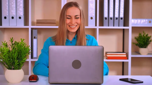 近代的なオフィスに机に座って陽気な労働者 幸せな笑顔で画面をコンピューターを使用してブロンドの髪と魅力的な若いビジネス女性 従業員の身に着けている明るいシャツ — ストック写真