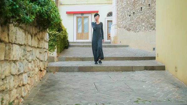 在老城的大街上走楼梯的女孩 女性使用手机的应用程序 她穿着休闲的灰色长裙 混合种族模型快乐和微笑 夏季游客在地中海城市漫步 — 图库照片