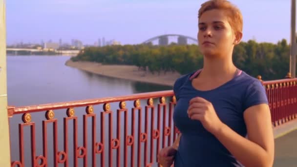 白人女性アスリート橋スローモーション女性でブロンドの髪の列車とで実行されている早朝の美しい青い空と川に表示します — ストック動画