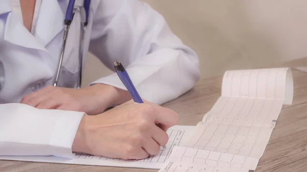 O médico olha cardiograma e faz anotações em um documento . — Fotografia de Stock