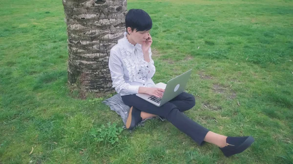 Frau sitzt unter Baum im Park und arbeitet — Stockfoto