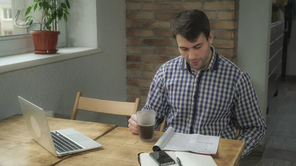 Привлекательный мужчина пьет кофе и читает газету, на столе телефон и ноутбук . — стоковое фото