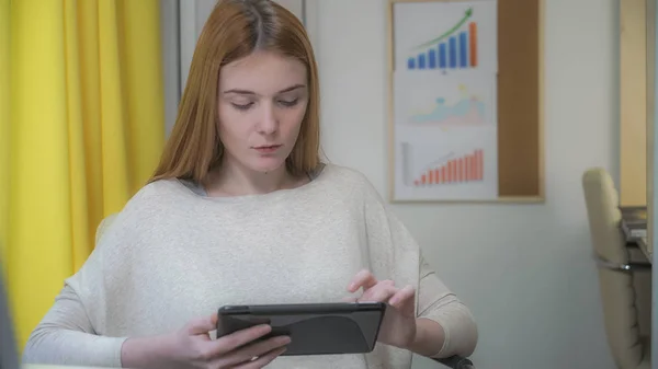 La mujer se sienta en el lugar de trabajo, en las manos sosteniendo la tableta . — Foto de Stock