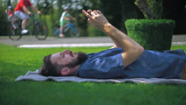 一个留着胡子躺在公园草地上的毯子上的年轻人 电话里写着一条信息 他笑了 下一个路过的骑自行车的人 — 图库照片