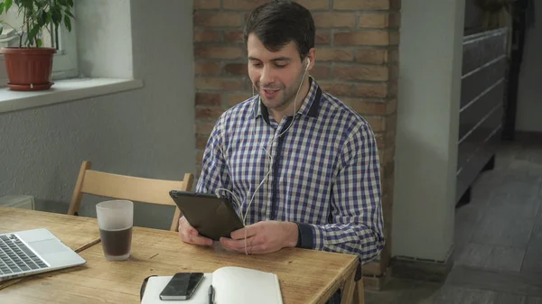 Мужчина сидит за столом с планшетом, наушниками в ушах, разговаривает онлайн . — стоковое фото