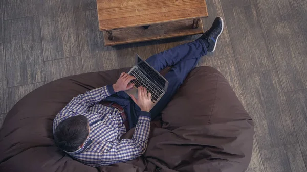 Un homme allongé dans un fauteuil, tenant un ordinateur portable et tapant . — Photo