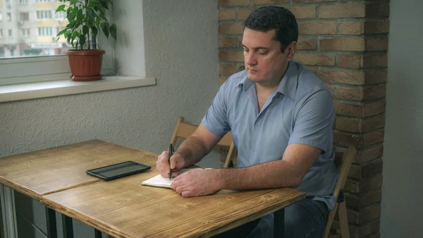 Дорослий чоловік сидить у кафе з щоденником і планшетом, роблячи нотатки в блокноті . — стокове фото