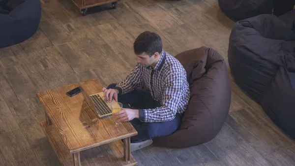L'homme est assis sur une chaise douce et tapant sur un ordinateur portable. Sur la table se trouve un verre de jus d'orange . — Photo