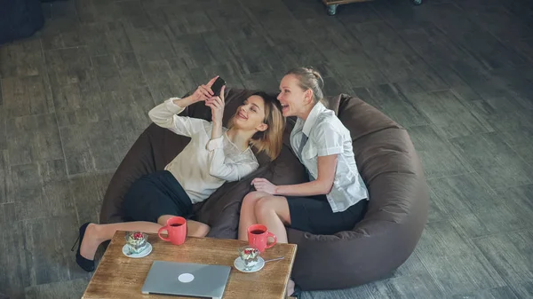 Две увлекательные блондинки, лежащие на диване и просматривающие фотографии по телефону, обсуждающие быстро сделанные селфи, громко смеющиеся . — стоковое фото