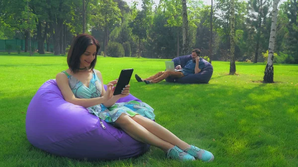 Chica morena joven está sentado en una silla en el parque y deslizar páginas en su tableta . — Foto de Stock