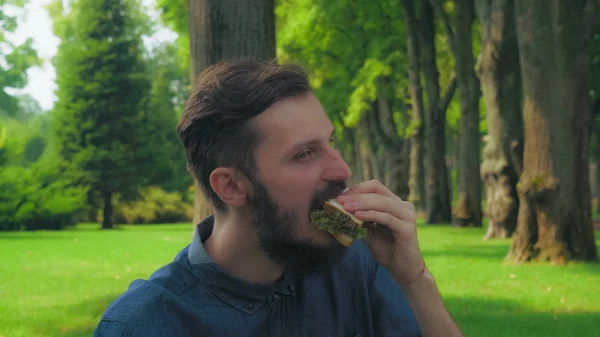 Az ember eszik saláta egy szendvicset a parkban. Élvezik az íze, és becsukja a szemét. — Stock Fotó